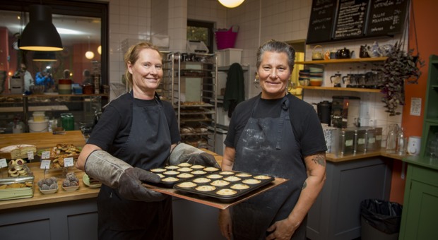 Två kvinnor håller upp ugnsplåtar med bullar i sitt bageri.